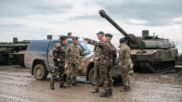 Devant le pick-up bleu à sérigraphie rouge et blanche des néeerlandais, et les chars de l'Armée française, 2 prévôts et trois gendarmes néerlandais échangent. L'un des prévos désigne du bras quelque chose vers la gauche à un gendarme néerlandais