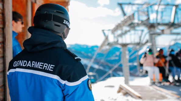 Vue de dos d'un gendarme en parka bleue, porteur d'un casque de montagne , devant les remontées mécaniques et la montagne en flou. A gauche, un chalet avec une personne dont on aperçoit en flou le vsage de profil avec des lunettes et un haut bleu clair