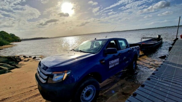 Sur une plage de sable jaune, un véhicule 4x4 bleu marine, portant la mention "Gendarmerie - Brigade fluviale" sur la portière tracte une barque flottant dans l'eau. Sur la droite de l'image, apparait une passerelle faisant la jonction entre l'eau et la plage. Dans le ciel est bleu et légèrement laiteux, apparaissent des nuages blancs et un solein voilé.