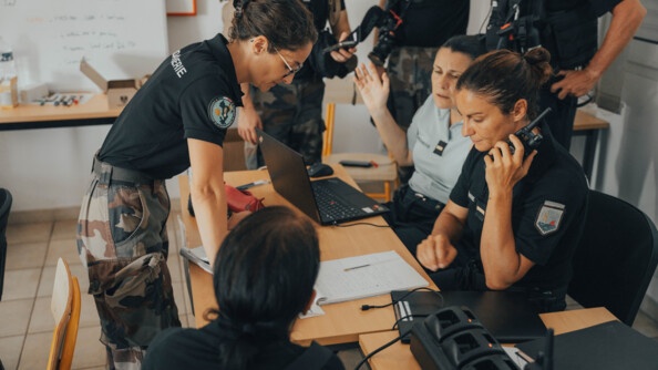 Plusieurs femmes gendarmes sont réunies autour d'une table sur laquelle sont posés un ordinateur et des cahiers. L'une d'elle a un talkie walkie à la main.