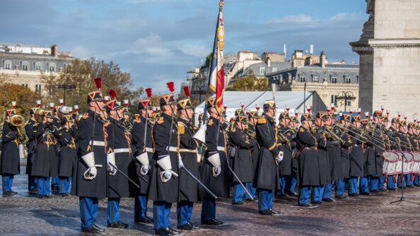 Des hommes, nombreux, de la Garde républicaine se tiennent debout sur un sol pavé. L'un d'eux rient un drapeau, tandis que d'autres tiennent leur instrument de musique (tambours, trompettes).Derrière eux, on aperçoit les façades d'imeubles en pierre. Le ciel est gris et bleu par endroits.
