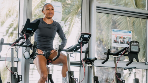Une sportive vêtue d'une tenue grise, pédale, souriante, sur un vélo fixe. Ses mains sont posées sur le haut de ses cuisses.