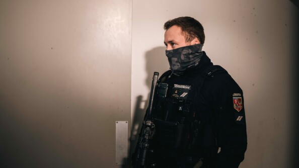 Un gendarme se tient debout, de biais par rapport &agrave; l'objectif, dans un lieu sombre. Il tient un fusil contre son corps. Son visage est partiellement recouvert d'un cache-nez.