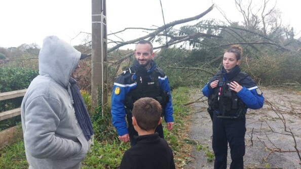 Un homme et une femme gendarmes, en tenue, debout, de face, sur la photo, disucutent avec un homme que l'on voit de biais, emitouflé dans un sweat à capucje gris, avec à ses côtés, un jeune garçon, qui se tient dos au photographe. Derrière eux, on aperçoit le ciel gros et des arbres couchés après la tempête, ainsi qu'un poteau électrique