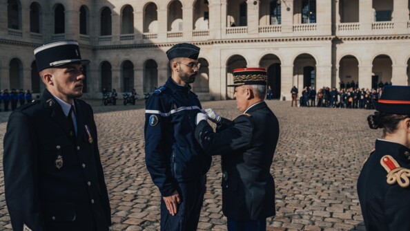 Le directeur général de la gendarmerie nationale remet une médaille à un gendarme adjoint volontaire dans la cour pavée des invalides. A gauche un gendarme en vareuse, à droite, le dos d'une garde républicaine. Autour de la cour, d'autres gendarmes et des civils