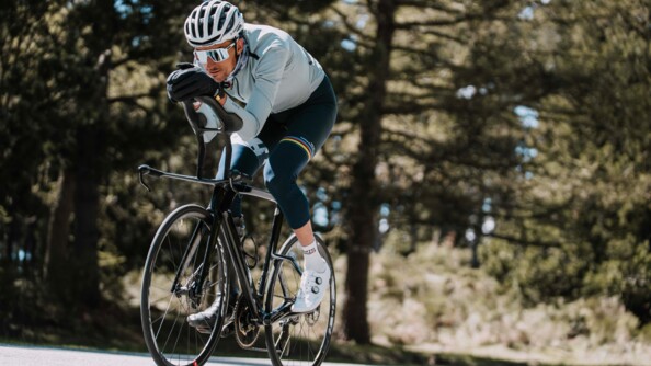 Un cycliste avance sur son v&eacute;lo, le corps pench&eacute; en avant, gravissant une c&ocirc;te. V&ecirc;tu d'une tenue bleue, il porte un casque, des lunettes de soleil blanches et des gants noirs.