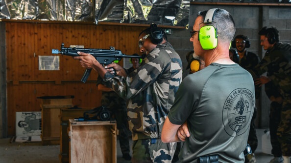 Un volleyeur du CVB52 en treillis kaki, vu de profil, en train de tirer au fusil mitrailleur, sous le regard d'un instructeur de l'école de Chaumont, en tee-shirt kaki, vu de dos.  Dans le fond, à droite, d'autres volleyeurs en treillis observent la scène.