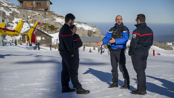 Un gendarme discute avec trois pompiers sur la neige à côté d'une station . A gauche la queue d'un hélicoptère de la sécurité civile