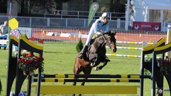 Une femme en tenue de gendarmerie sur un cheval en train de sauter un obstacle peint en jaune en noir