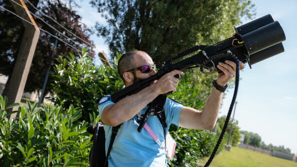 Un gendarme de profil, plan en buste, vêtu d'un polo bleu barré par une lanière rose portant la mention "Paris 2024", son accréditation est sur le côté, sous son bras. Il porte des lunettes de soleil, un sac noir sur le dos et pointe un fusil de lutte anti-drone vers le cile. En arrière-plan, des buissons, des arbres, un pré.