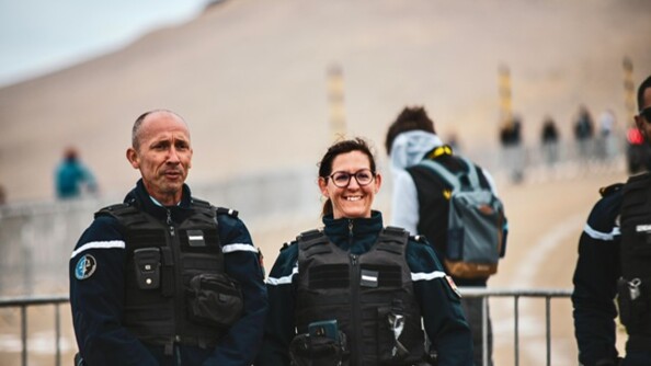 Deux gendarmes de face, souriants, et un troisième à droite, dont on ne voit que la moitié, sont debout devant une barrière. Derrière eux on devine des gens à pied et le paysage minéral du Mont Ventoux.