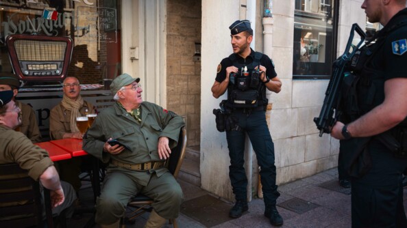 Deux gendarmes échangent dans la rue avec des hommes, habillés en tenue militaire pour l'occasion et assis en terrasse.
