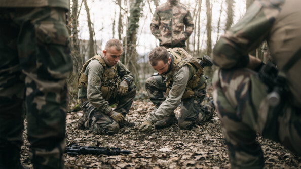 Deux militaires en treillis au sol expliquent une manoeuvre. Quatre militaires en treillis debout les observent
