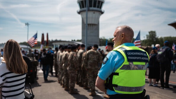 Un gendarme des transports aériens de dos, regardant vers la gauche. AU second plan de nombreux militaires américains alignés. Une foule est présente sur le tarmac