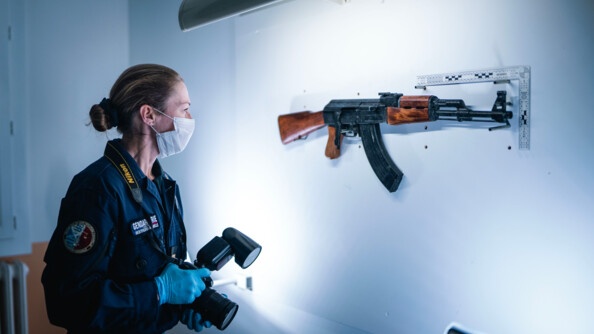 Une femme gendarme, équipée de gants bleus, d'un masque et tenant un appareil photo dans les mains, se tient face à un fusil, supendu sur un mur blanc, et éclairé par une lampe murale située au dessus