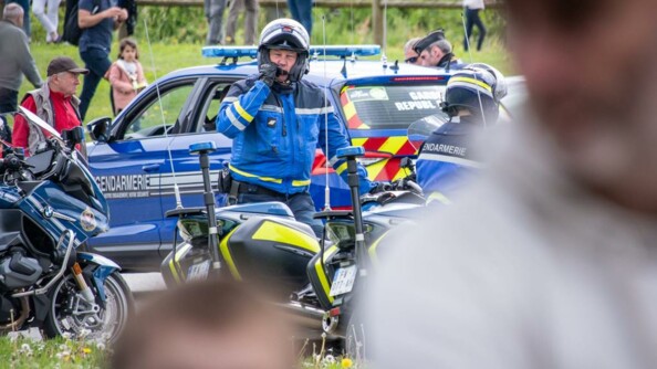 Deux personnages flous au premier plan. Au second plan, deux motocyclistes d'EDSR, l'un de face, debout à côté de sa moto, en train de parler dans son micro ; le second, vu de dos, sur sa moto. En arrière plan, une moto et une voiture gendarmerie et du public.