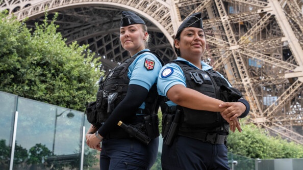 Deux femmes gendarmes  se tiennent debout, côte à côte, devant la Tour Eiffel. Toutes deux sont équipées d'un gilet pare-balles, porté sur leur polo bleu ciel.