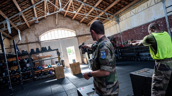 Dans un hangar de crossfit, de hommes de dos en treillis, dont celui de gauche et surmonté d'une chasuble jaune fluo, tirent au pistolet laser sur des cibles carées posées sur des caisses en bois