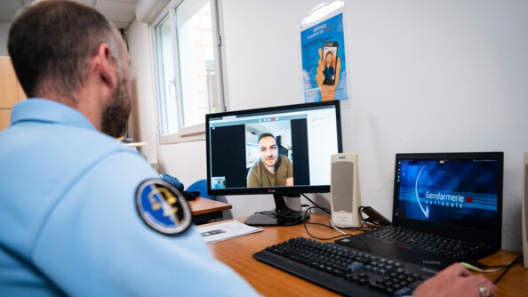 Un gendarme, de dos, à gauche, prend la plainte d'une victime en visio-conférence, dont on voit le visage sur un écran d'ordinateur posé sur le bureau.