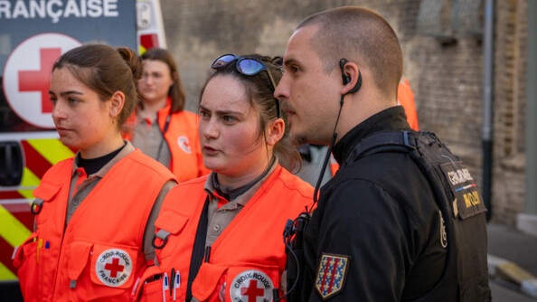 Photo d'un gendarme, aux c&ocirc;t&eacute;s de deux profesionnelles de la croix rouge fran&ccedil;aise. On aper&ccedil;oit le v&eacute;hicule de la Croix rouge en arri&egrave;re plan.