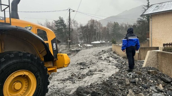 Au premier plan, à gauche, l'avant d'un engin de chantier jaune. Au second plan, à droite, un gendarme de dos, vêtu d'un blouson de pluie, capuche sur la tête, sur une digue de fortune érigée à proximité d'un fleuve de boue dévalant au milieu des habitations.