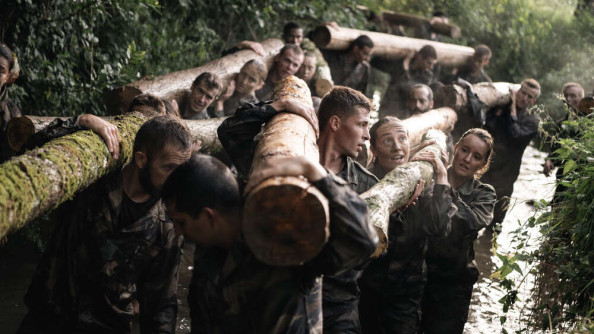 Des élèves gendarmes en treillis progressent dans un cours d'eau en transportant des troncs d'arbre en binôme.