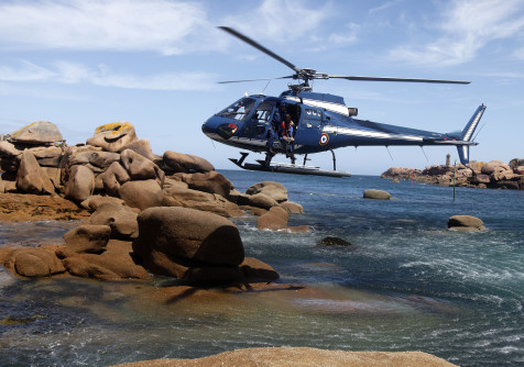 Un hélicoptère bleu marine de la Gendarmerie nationale est en vol stationnaire au-dessus d’un littoral rocheux. Un plongeur en tenue de sauvetage est positionné à la porte latérale, prêt à intervenir. Le paysage est constitué de gros rochers arrondis, partiellement immergés, et d’une mer calme sous un ciel bleu légèrement nuageux. À l’arrière-plan, un phare est visible sur un îlot rocheux.