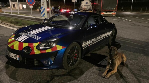 A gauche, un véhicule rapide d'intervention (VRI) Alpine 110 de la gendarmerie, à droite, sagement couché, un chien d'une équipe cynophile. En arrière plan, les gendarmes contrôlent des véhicules et des poids lourds.