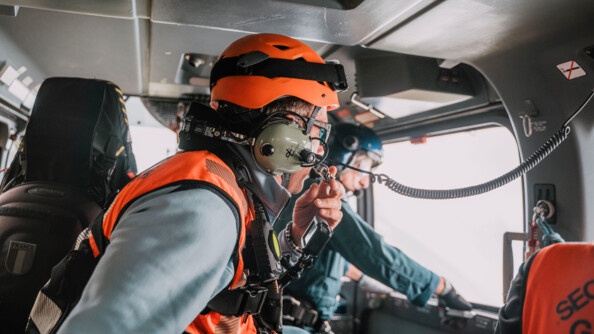 Vue de l'intérieur d'un cockpit d'hélicoptère gendarmerie, deux gendarmes sont un vếtu d'une chasuble orange. Au fond l'autre, flou, en combinaison de vol, s'apprête à ouvrir la porte latérale