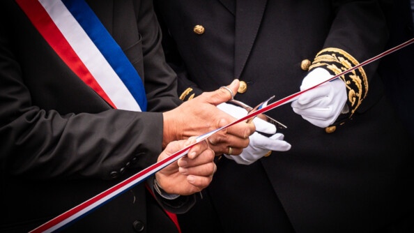 Photo représentant les mains de deux personnes, dont l'une porte des gants blancs, et coupent ensemble un ruban tricolore aux couleurs de la France. l'une des personnes porte un uniforme noir, dont le bout des manches sont gansées de broderies dorées, tandis que la seconde (un élu), porte un costume noir et une écharpe tricolore sur la poitrine.