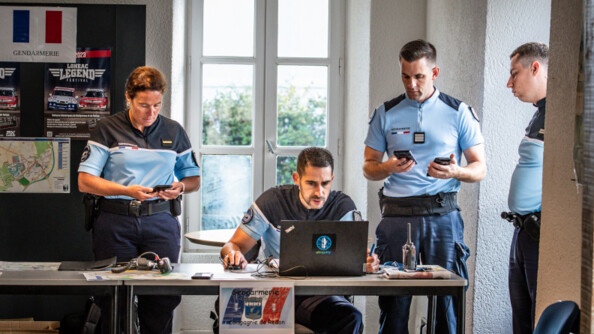 Derrière une table, dans une salle avec une fenêtre, 4 gendarmes, trois hommes et une femmme à gauche. Un des hommes est assis derrire un ordinateur portable posé sur la table. Les deux autres gendarmes sont à droite et regardent vers l'écran