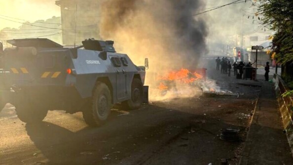 Un véhicule blindé de la gendarmerie, de couleur bleue, circule sur une route, se dirigeant droit sur une barricade en feu barrant la voie, dont s'échappe une épaisse fumée noire montant en dircetion du ciel.