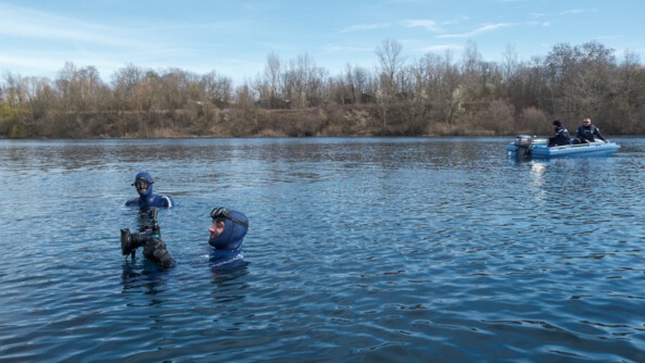 Plan large du plan d'eau sur lequel l'on voit en fond, l'un des bateaux de la gendarmerie et en premier plan, deux plongeurs de la gendarmerie entrain de prendre des photos et autres relevées d'enquête.