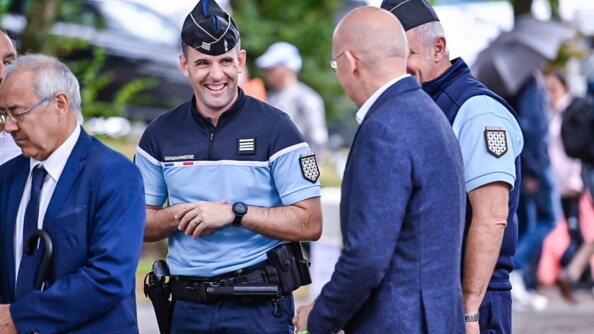 Un commandant de gendarmerie se tient les mains en riant, regardant un second homme vers la droite, en cosume bleu marine. Derrière ce dernier, un second gendarme et à gauche, un autre homme en costume