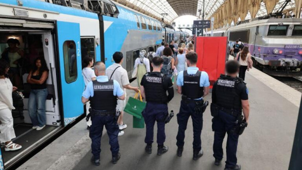 Photo de qautre gendarmes, de dos, debout côte à côte sur le quai d'une gare. Un train bleu et blanc, dont les portes sont ouvertes, est  stationné en gare.