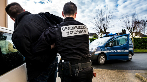 Au premier plan à gauche, de dos, un gendarme interpellant un homme vêtu d'un blouson noir contre une voiture blanche. En arrière plan, un Patrner gendarmerie, portière conducteur ouverte