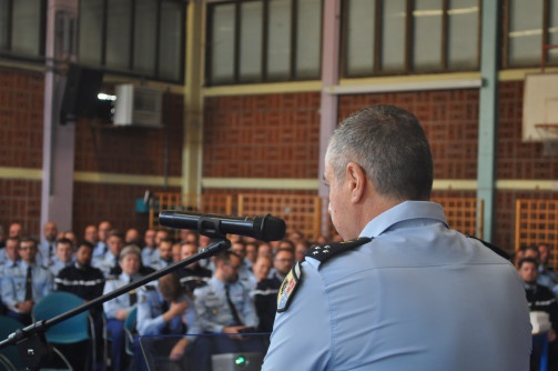 La photo représente un gendarme, de dos, s'exprimant à un micro, face à une assemblée de gendarmes.