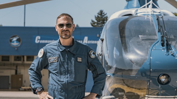 La photo représente un pilote d'hélicoptère, posant debout, mains sur les hanches, devant l'hélicoptère bleu de la gendarmerie.