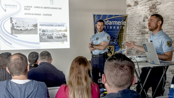 Poursuite de la présentation par deux gendarmes debout dans une salle, sur la droite, aux personnes assises sur des chaises. Le power point projeté indique "partageons la route"