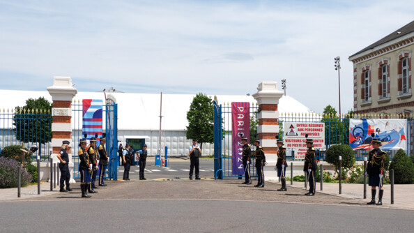 De part et d'autre d'une grille d'entrée bleue aux couleurs des Jeux de  Paris 2024, huit élèves-officiers de la gendarmerie nationale en tenue de tradition. Au centre de la grille, trois gendarmes en tenue d'intervention