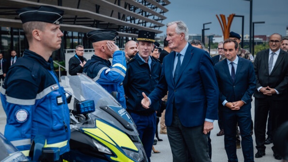 Le premier ministre, Michel Barnier, tend la main vers un gendarme en faction devant le stade de France (Saint-Denis). D'autres personnalités officielles se tiennent à leurs côtés.