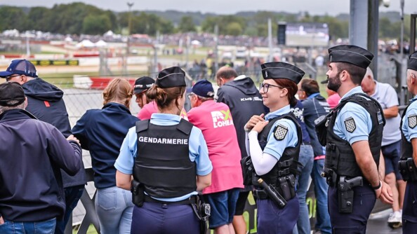 Une gendarme de dos et deux de profil, sur la droite de la première. Derrière eux, le public accoudé aux barrières de course. EN arrière-plan, le village de course, flou
