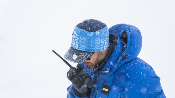 Dans un paysage enneigé, un homme vếtu d'un anorak et d'une casquette de couleur bleue, dont une aperçoit uniquement le haut du corps, se tient debout, la tête inclinée vers l'avant. Il tient dans sa main droite un talkie walkie