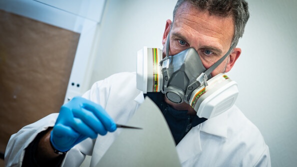 Un homme en blouse blanche, en buste, portant un masque avec filtres, tient de la main droite gantée de bleu une pince à épiler tenant une feuille blanche