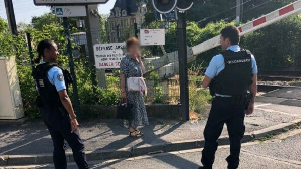 Un gendarme vu de profil et un gendarme vu de dos dispensent des conseils de pr&eacute;vention &agrave; une passante, &agrave; proximit&eacute; d'un passage &agrave; niveau, dont les barri&egrave;res sont en train de descendre.