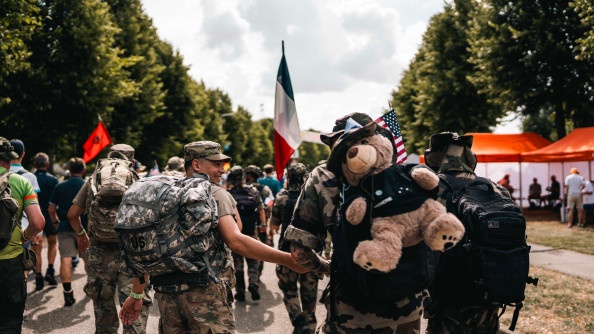 Plussieurs militaires en tenue kakie, marchent ensemble. Ils sont photographiés de dos. Un drapeau français émerge du groupe. Un gros ours en peluche est accroché au sac de l'un des militaires.