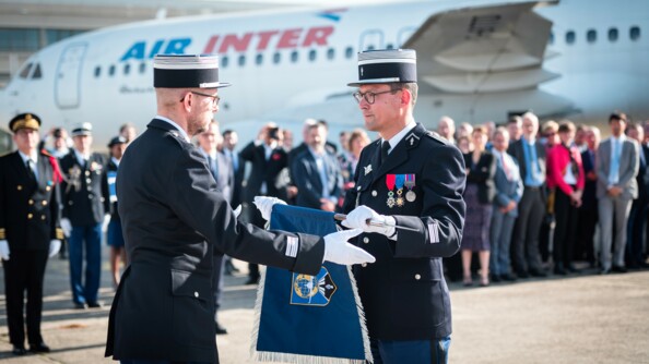 Au premier plan, le colonel Philippe Mirabaud, à droite, reçoit le fanion de la GTA des mains de son précédesseur, lors de sa prise de commandement, à l'été 2022. Au second plan, un parterre de personnes, civiles et militaires, et un avion sur le tarmac.