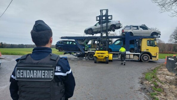 Au premier plan, un gendarme de dos. Au second plan, un chariot élévateur jaune charge des épaves de véhicules sur un camion à tête jaune.