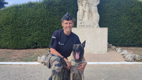 La photo représente une femme gendarme, souriante, portant un pantalon kaki, un polo et un calot noirs. La militaire est accroupie aux côtés de son chien, qu'elle tient contre elle. Derrière eux se dresse une statue blanche sur laquelle est inscrit "Toi et moi, pour eux".