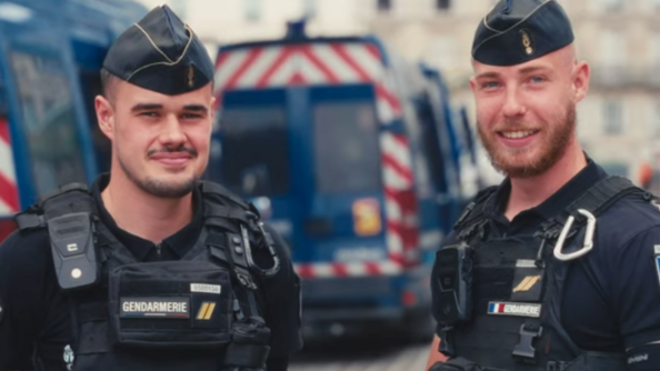 Deux gendarmes en tenue se tiennent côte à côte, souriants, devant les fourgons de lagendarmerie